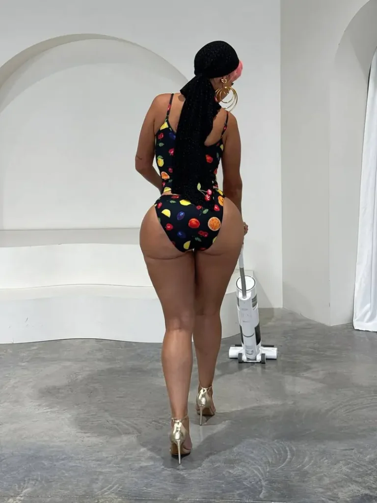 Amirah Dyme vacuuming a room with her back to the camera wearing a navy one-piece swimsuit with orange, strawberry and lemon prints, gold high heels, a black headscarf and large gold hoop earrings with short pink hair